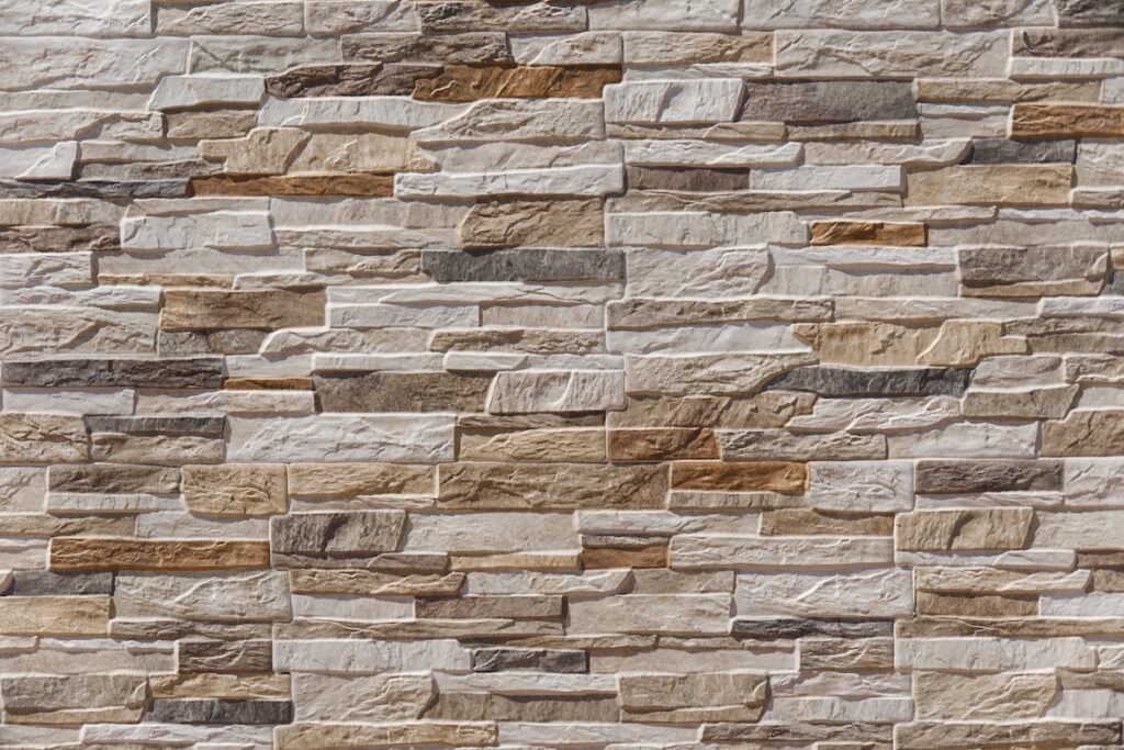 Full frame texture shot of a modern decorative stone wall built with beige, brown, and grey slate tiles. Architectural background showing stacked stone masonry, clean surface, and outdoor sunlight.