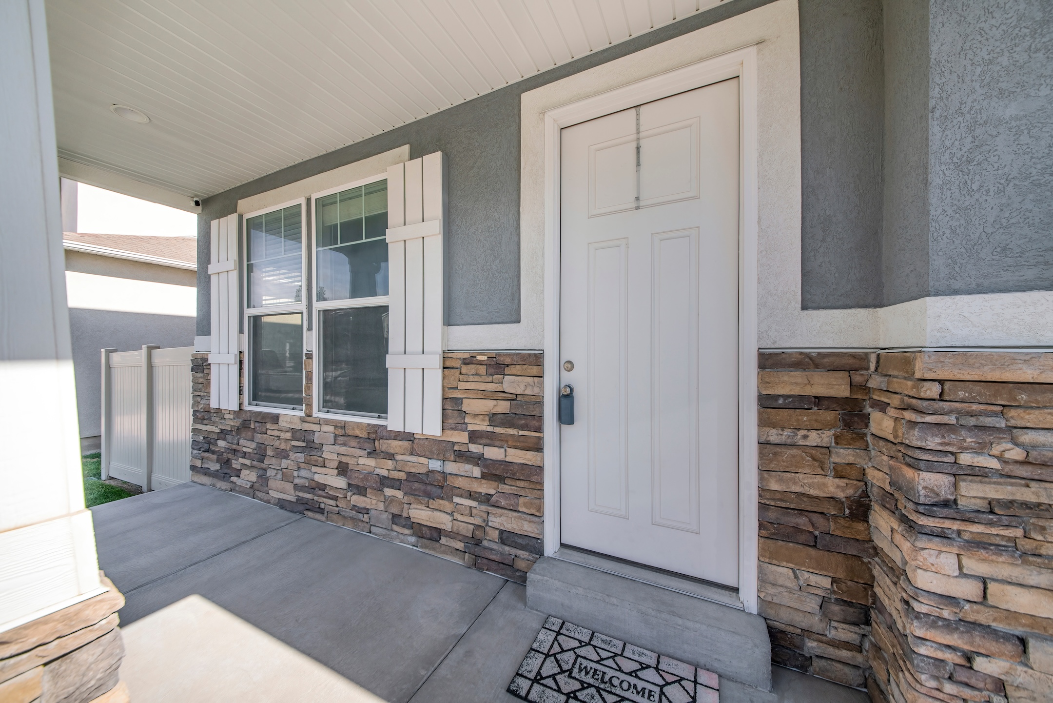 Exterior of a house with gray and stone veneer siding. There is a white door with lockbox near the windows with white fake shutter design near the vinyl white fence on the side.